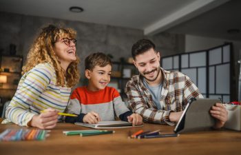 A woman, boy, and man sit at a table with art supplies, smiling and looking at a tablet.